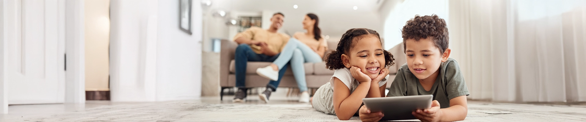 Image of young brother and sister playing on tablet device while father and mother observe in the background